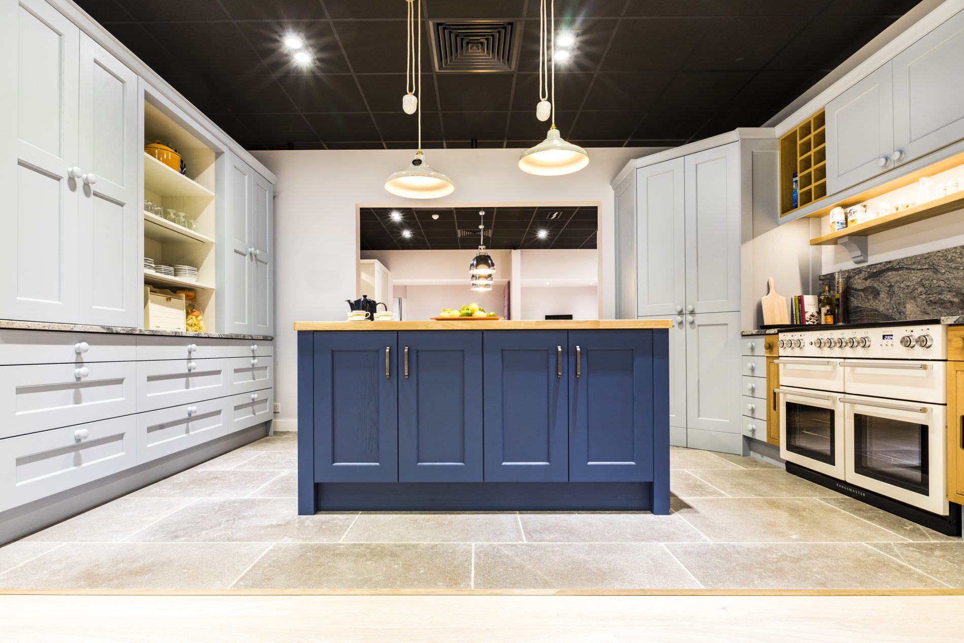 Panoramic shot of our kitchen showroom featuring our pantry blue kitchen, range oven, pendant lighting and limestone tiled flooring